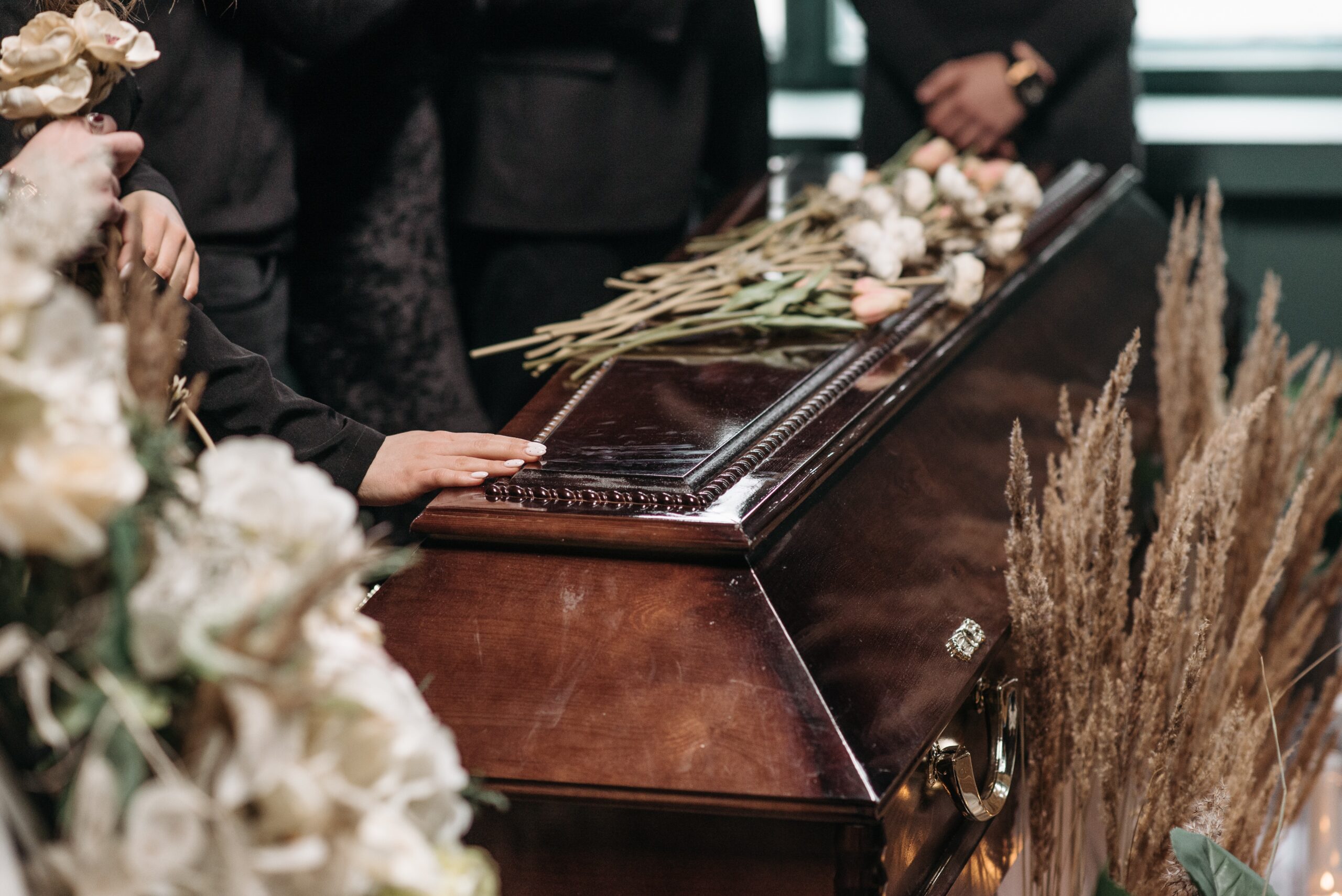 Brauner, glänzender Sarg in der Trauerhalle bei einer Beerdigung. Auf dem Sarg liegen Blumen und daneben stehen schwarz gekleidete Trauergäste